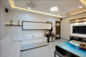 The Compact Living Room with White & Brown Combination has L Shaped White Upholstered Sofa with Wooden Side Table cum Storage Unit