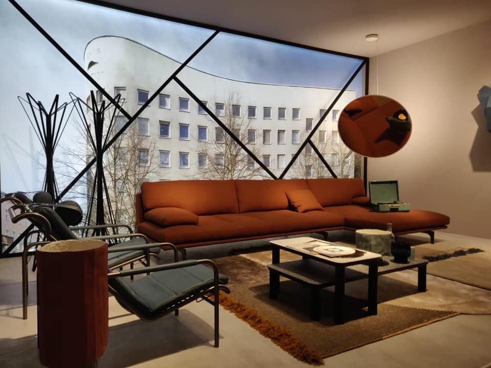 The Living Room with a Full Height Glass Facade in an Inclined Line Pattern Frame has Brick Red Sectional Sofa with Metal Legs